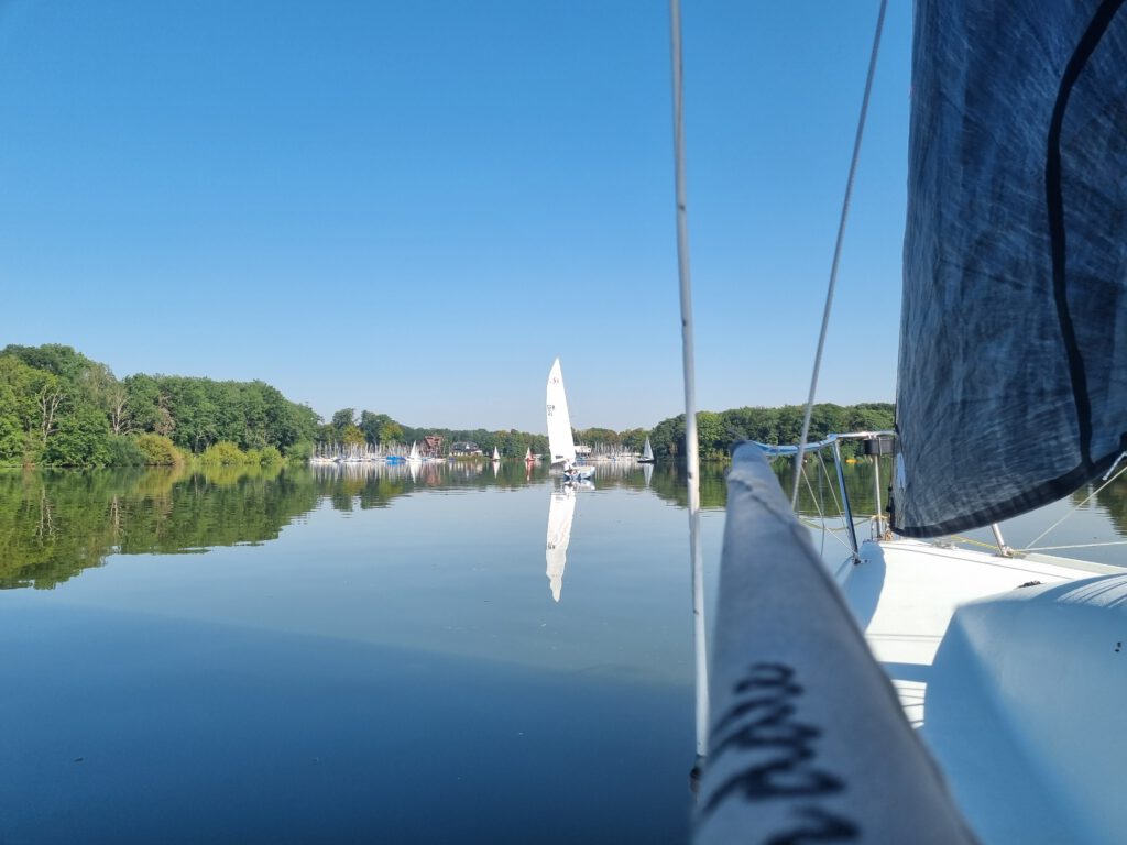 Blick von einem Segelboot auf einen See mit spiegelnder Oberfläche bei schönem Wetter. Im Hintergrund ist ein Segel eines anderen Bootes zu erkennen.